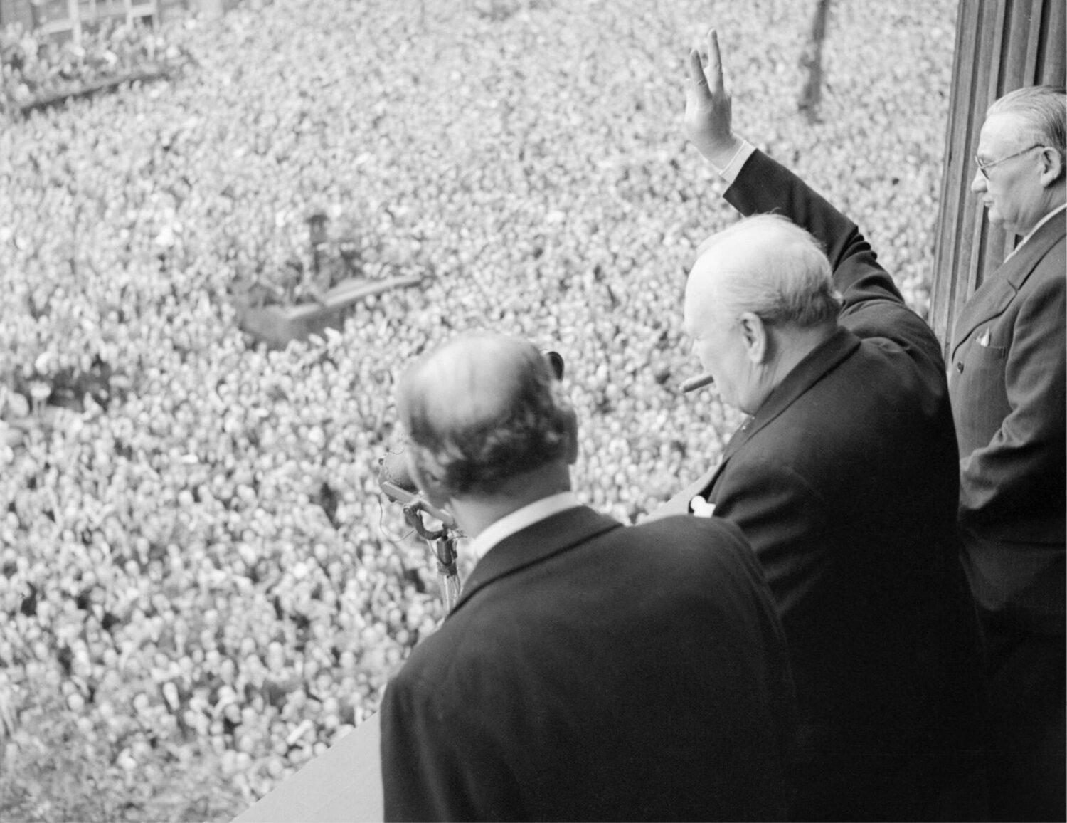 Winston Churchill waves to crowds in Whitehall, in London, as they celebrate VE Day, 8 May 1945. From the balcony of the Ministry of Health, Prime Minister Winston Churchill gives his famous 'V for Victory' sign to crowds in Whitehall on the day he broadcast to the nation that the war with Germany had been won, 8 May 1945 (VE Day). To Churchill's left is Sir John Anderson, the Chancellor of the Exchequer. To Churchill's right is Ernest Bevin, the Minister of Labour. (Public Domain)